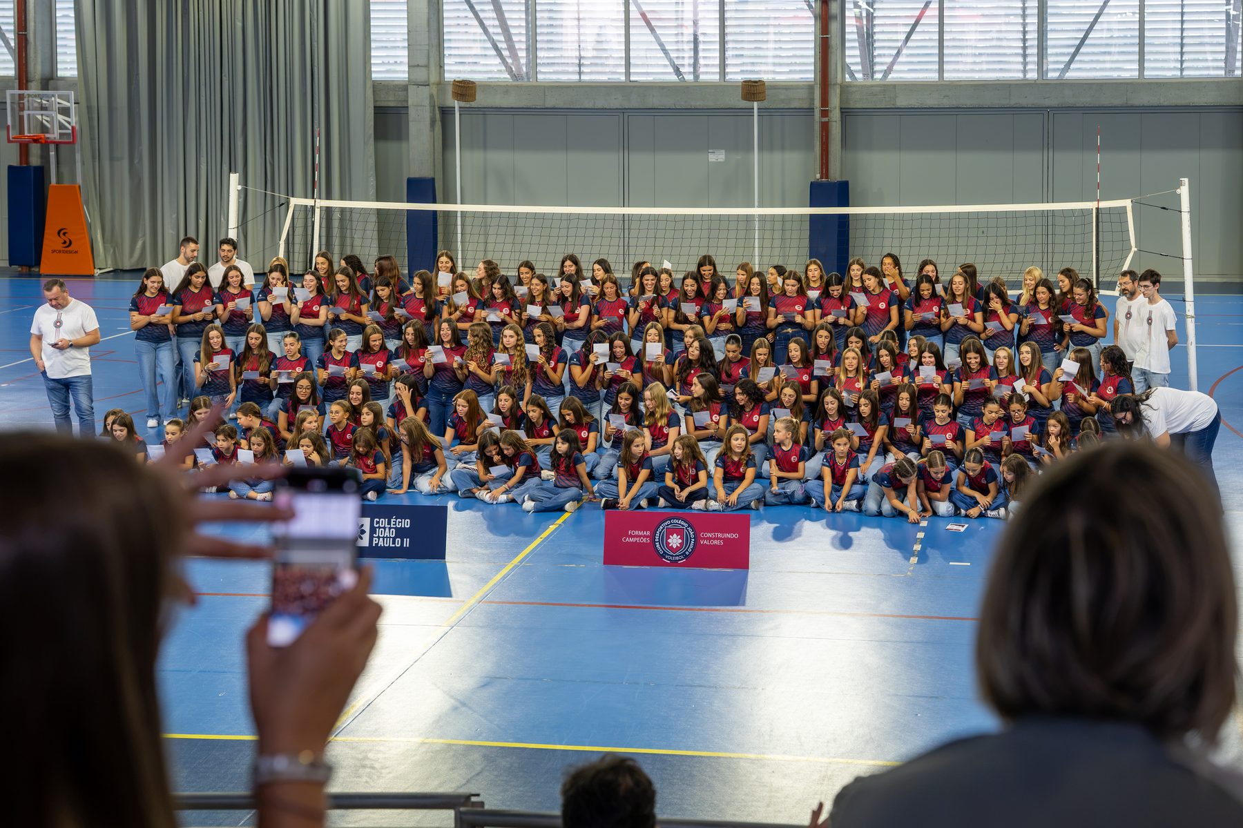 Todas as atletas do voleibol feminino reunidas no pavilhão, de pé e sentadas, a ler uma folha